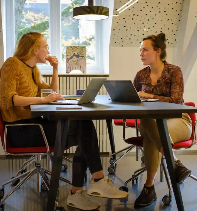 Twee collega's van Andersson Elffers Felix aan het praten aan een bureau met laptops over maatschappelijke vraagstukken